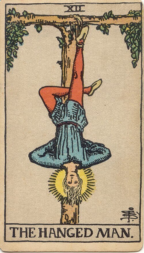 Tarot card showing a haloed man hanging upside down by one foot from a wooden frame, with his other leg bent behind him.